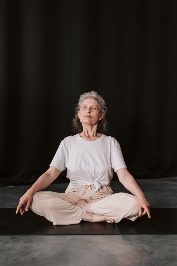 An elderly female yoga practitioner sits cross-legged on a black and reflective rectangular mat. 